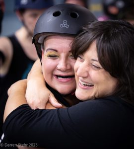 two people hugging and smiling, one wearing a roller derby helmet for safety